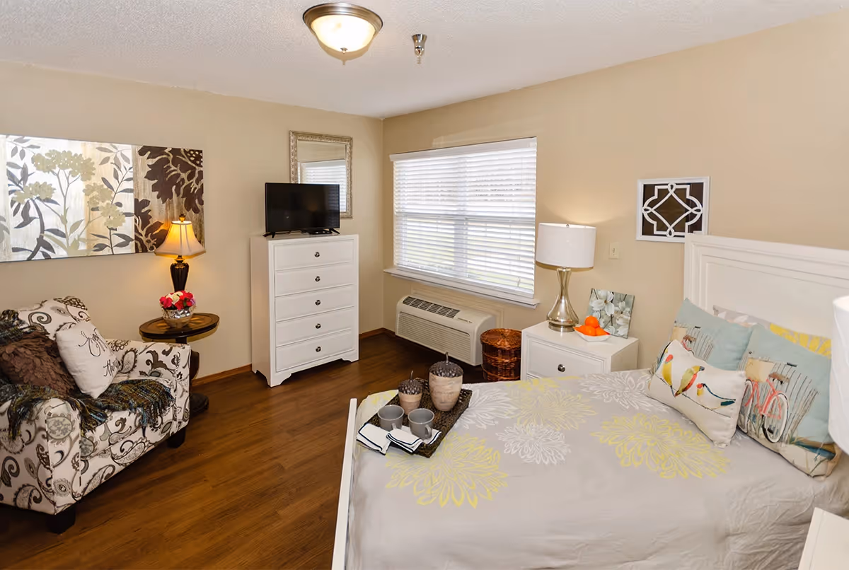 Well-lit furnished bedroom with a bed, patterned armchair, white dresser with a TV, nightstand, and a window with blinds.
