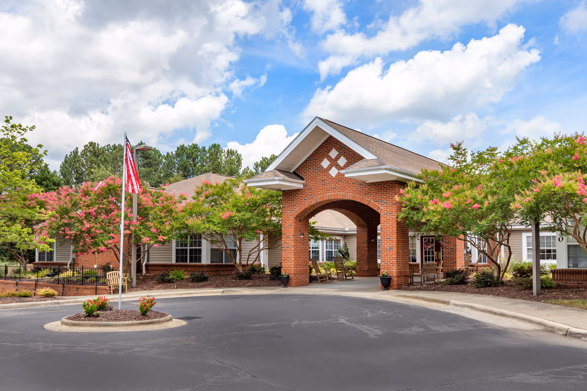 Front exterior view of a senior living facility with a brick entrance archway, rocking chairs under the arch, flowering trees, an American flag on a flagpole, and a clear blue sky with clouds.