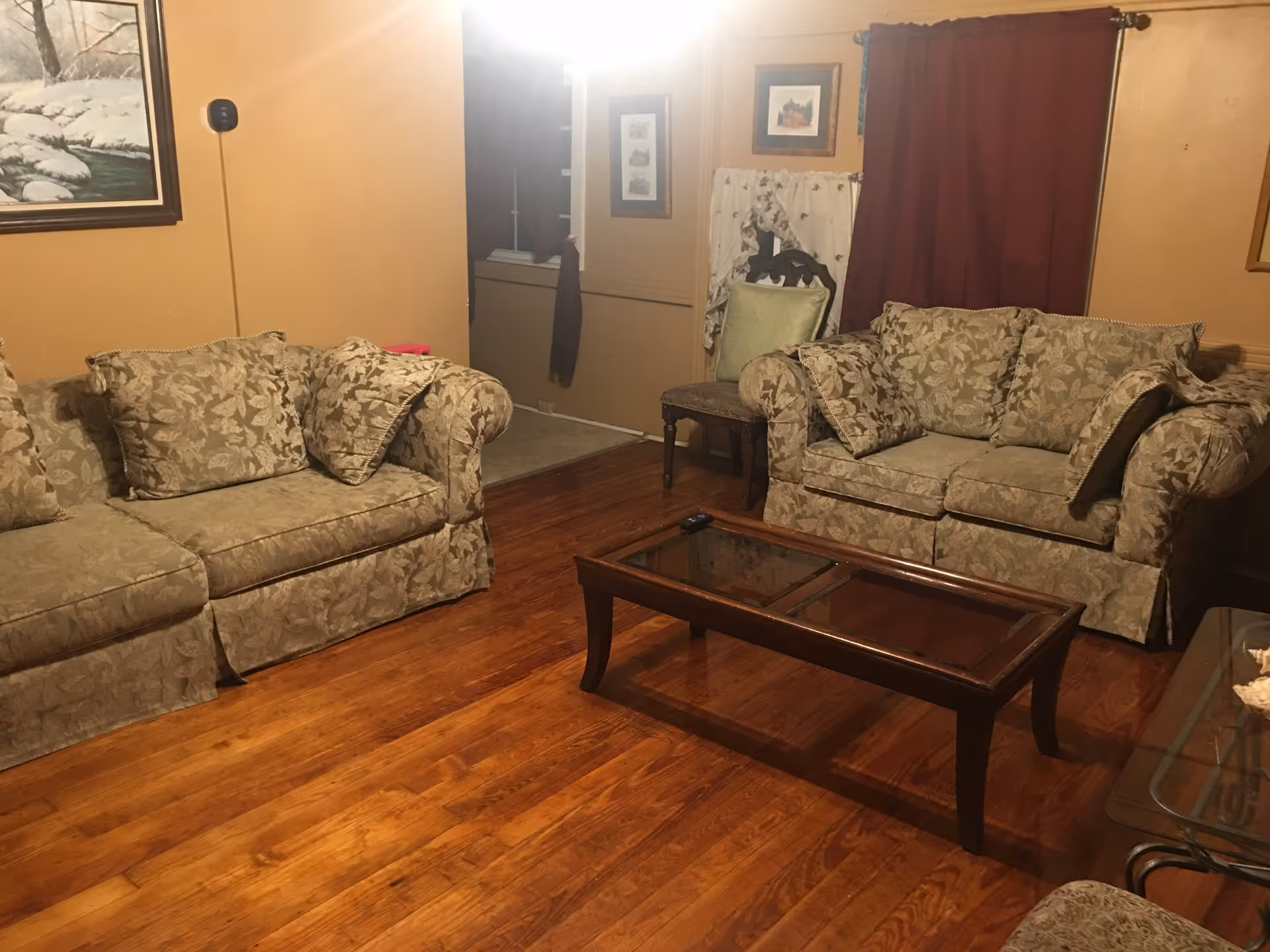 A cozy living room with two floral-patterned sofas facing each other, a wooden coffee table with a glass top in the center, hardwood floors, a chair with a green cushion against the wall, framed pictures on the walls, and a window covered by a dark red curtain.