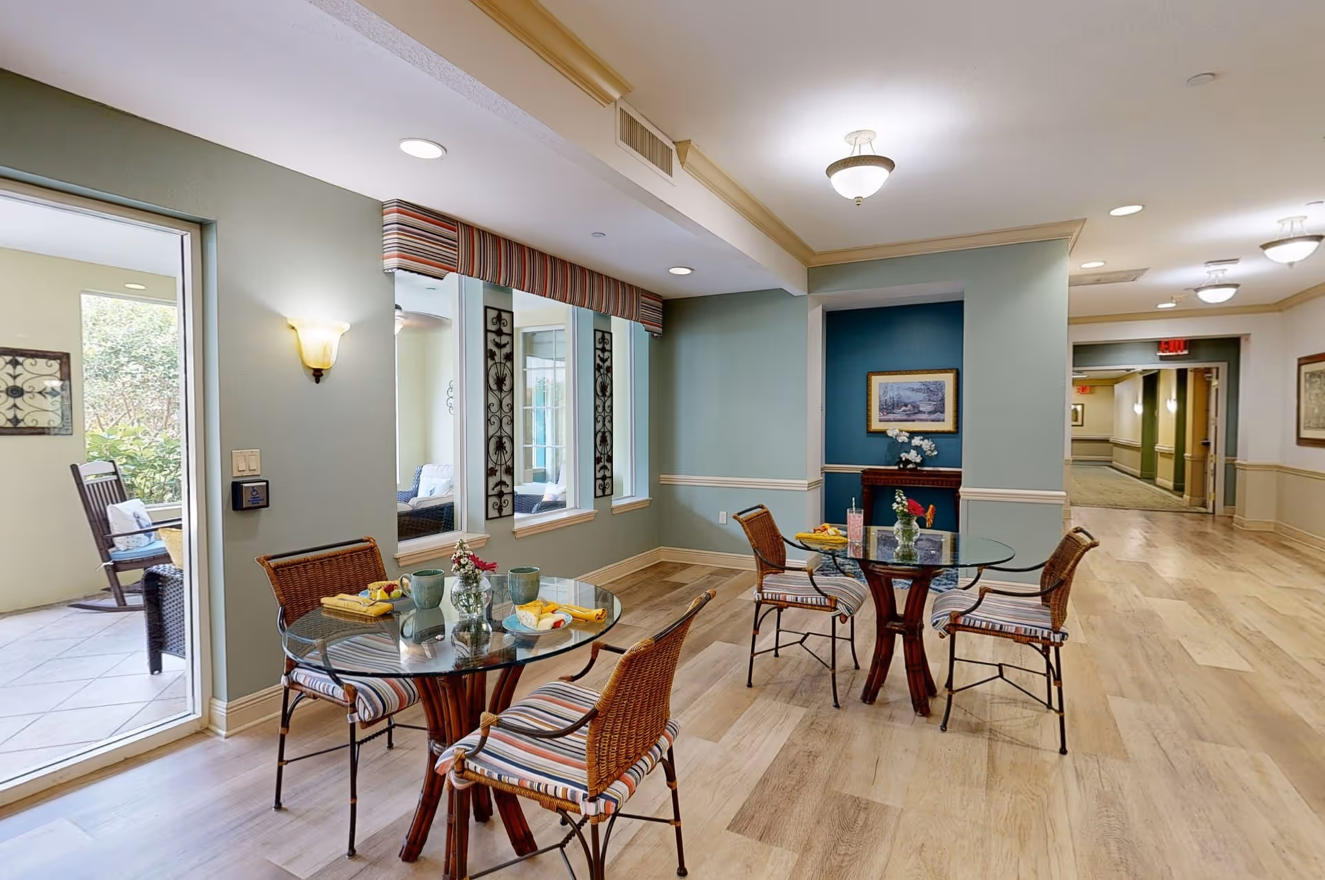 A bright and inviting common area with two round glass tables, each surrounded by four wicker chairs with striped cushions. The tables are set with plates of food, cups, and small flower vases. The walls are painted in soft green and blue tones with decorative wall art and a small alcove featuring a framed picture and flowers. Large windows and a door open to an outdoor patio area with rocking chairs. The floor is light wood, and ceiling lights provide warm illumination.