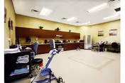 A spacious room with a kitchenette area featuring wooden cabinets, a microwave, and a refrigerator. There are chairs and tables arranged around the room, along with exercise equipment and neatly folded towels on a cart. The walls are painted light yellow and decorated with framed pictures.