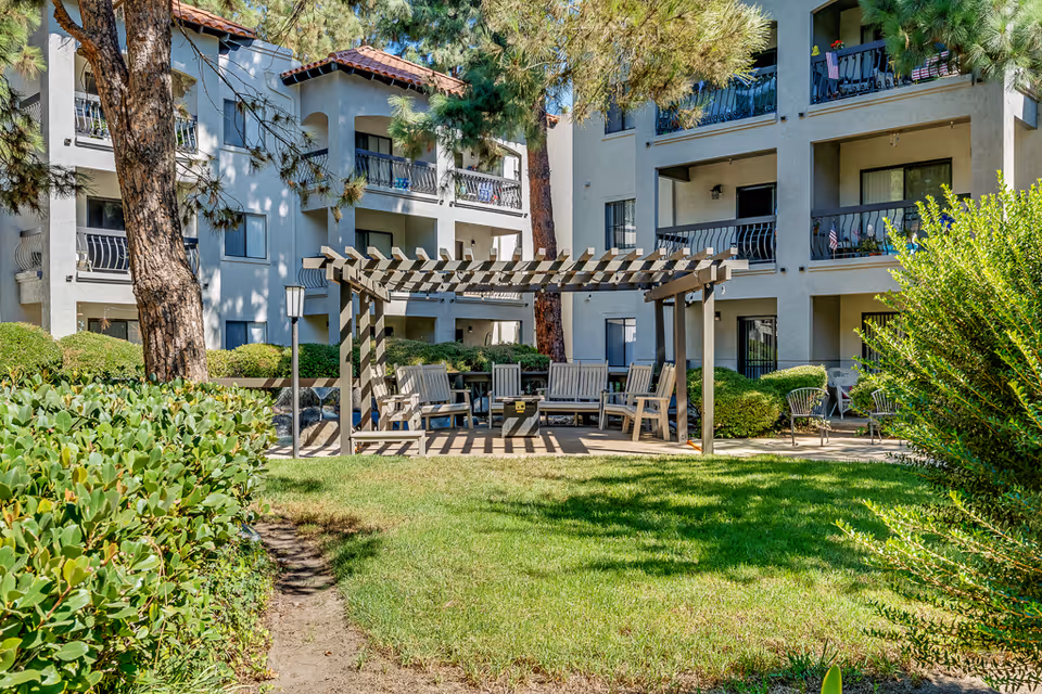 Courtyard with a wooden pergola, seating, lawn and landscaping in front of a multi-story senior living building with balconies.