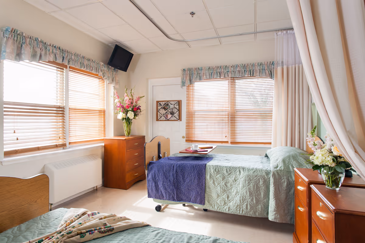 A bright nursing center bedroom with two beds, one with a green quilt and the other with a green quilt and a blue blanket. There are wooden dressers with flower vases, large windows with wooden blinds and floral valances, a wall-mounted TV in the corner, and a tray with a cup and plate on one bed. A privacy curtain is partially drawn on the right side.