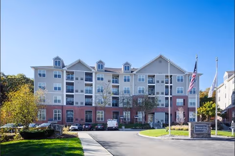 Front exterior view of a multi-story senior living facility building with a mix of brick and siding facade, several windows, and small dormer windows on the roof. There are parked cars in front, a driveway, a green lawn, trees, and flagpoles with the American flag and another flag near the entrance.