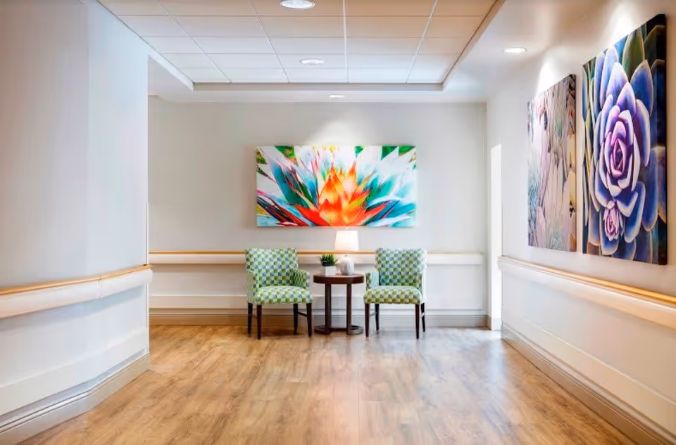 A bright hallway with light wood flooring and white walls featuring handrails. At the end of the hallway, there are two green patterned chairs with a small round table between them, holding a lamp and a small plant. Colorful floral artwork is displayed on the walls.