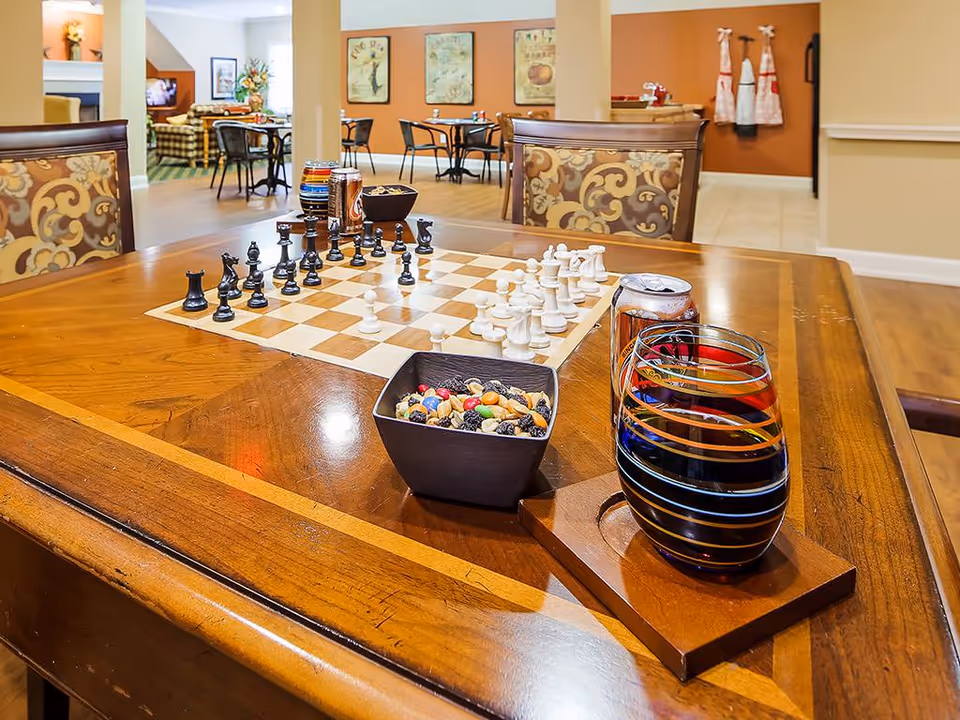 A wooden table with a chessboard set up for a game, a bowl of mixed nuts and candies, a colorful striped glass, and a soda can. In the background, there are chairs and tables in a warm-toned room with orange and beige walls, decorated with framed artwork and aprons hanging on the wall.
