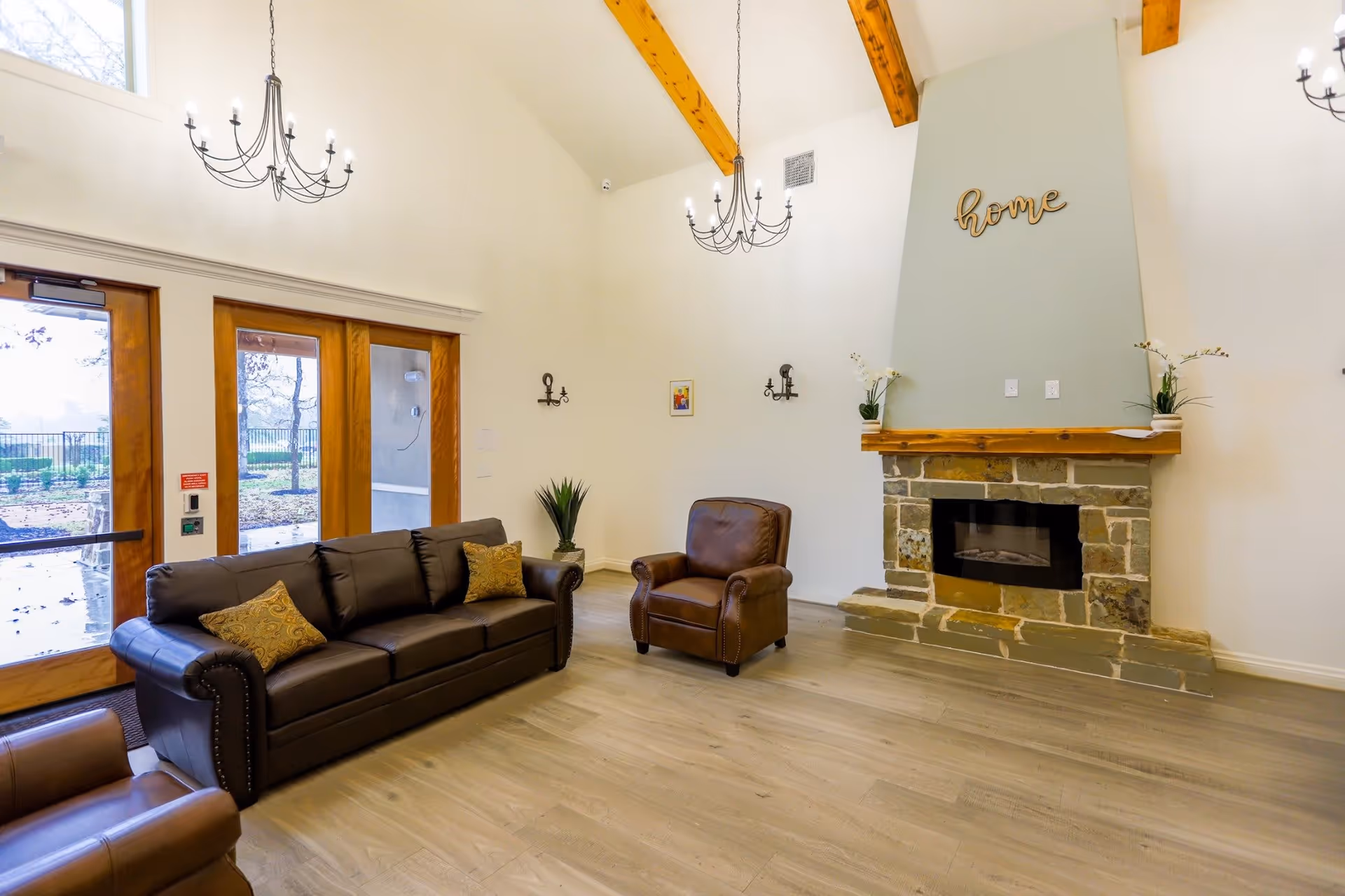 A bright and spacious living room with high vaulted ceilings featuring wooden beams. The room includes a dark brown leather sofa with two patterned cushions, a matching leather armchair, and a stone fireplace with a wooden mantel. Above the fireplace, the word 'home' is displayed in decorative wooden letters. Two chandeliers hang from the ceiling, and large wooden-framed glass doors lead outside to a garden area.