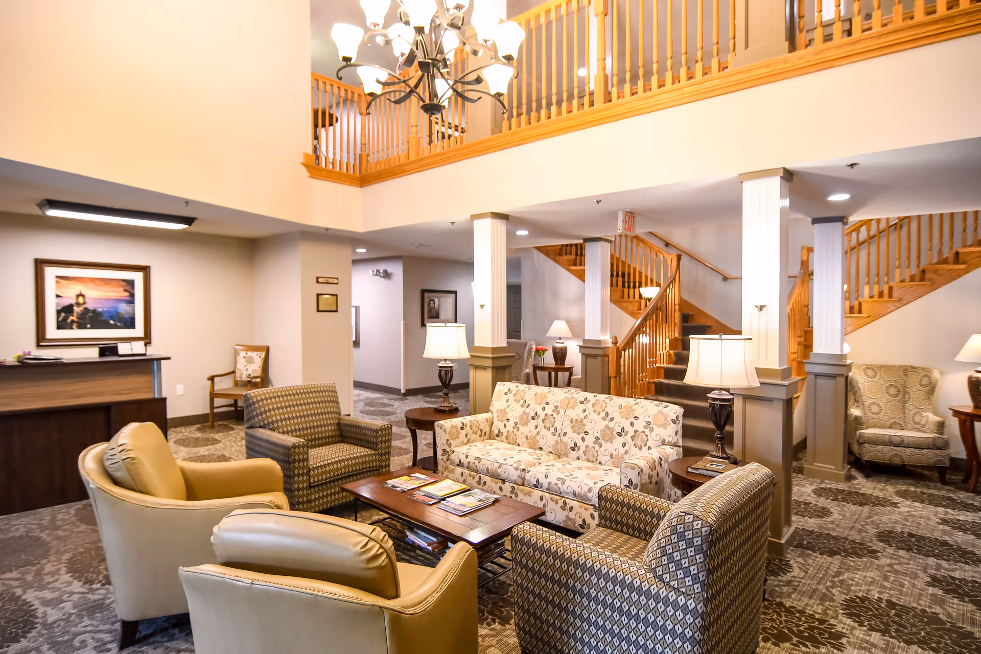 A welcoming senior living lounge with sofas and armchairs arranged around a coffee table, lamps, and a wooden staircase leading to an upper level.