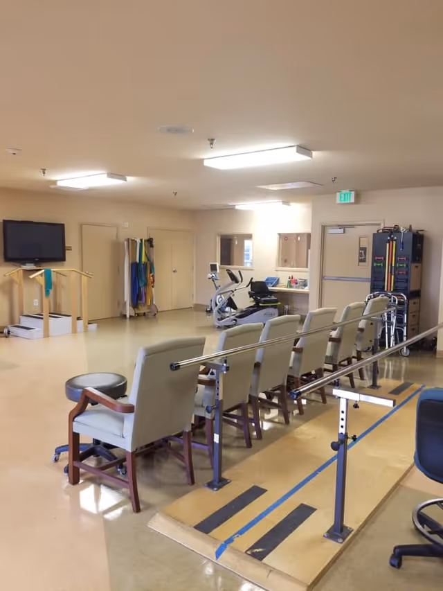 A rehabilitation room with parallel bars for walking exercises, several chairs lined up along the bars, a stationary exercise bike, a small set of stairs with handrails, and various exercise equipment and supplies in the background.