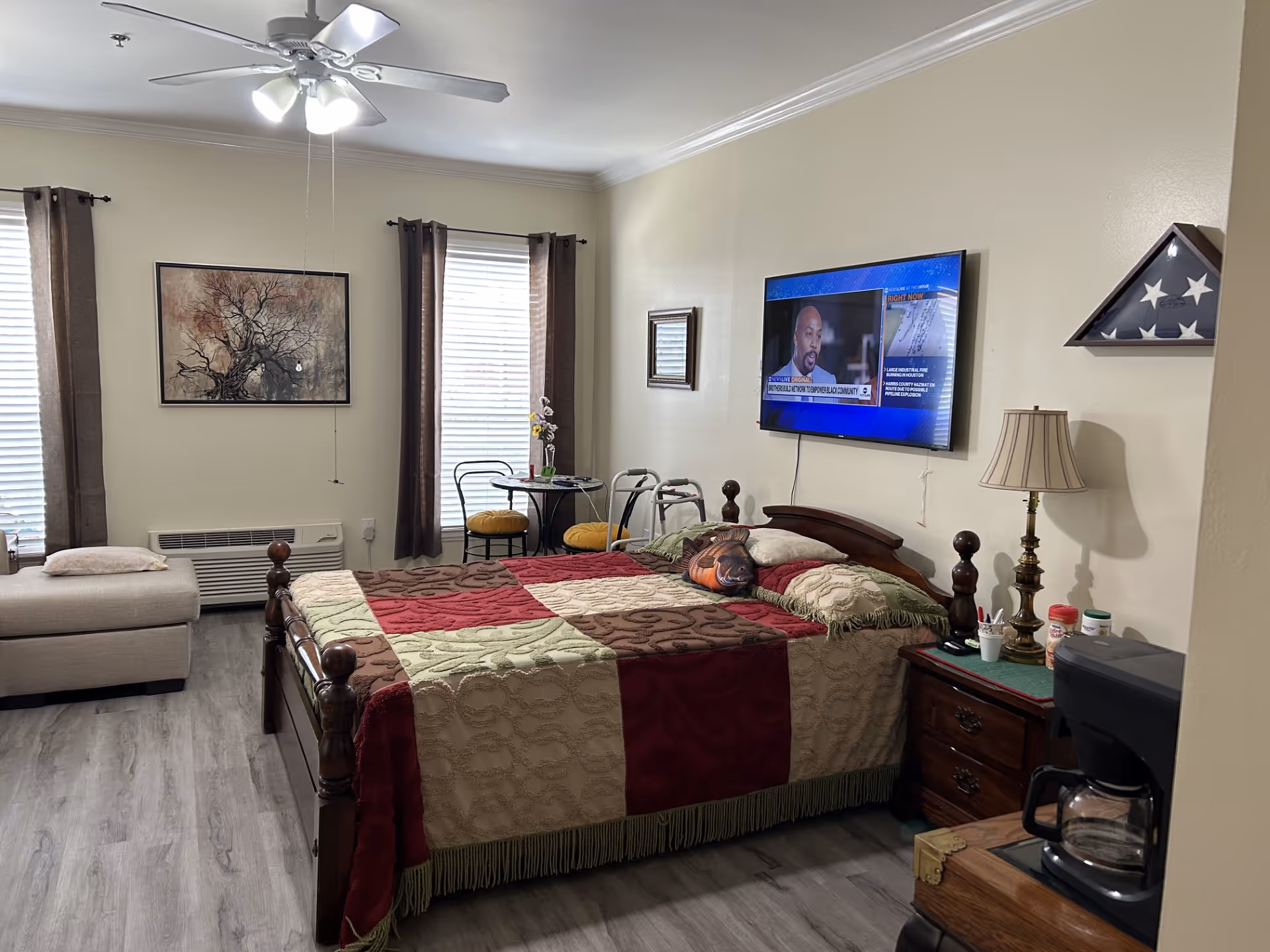 A senior living bedroom with a wooden bed covered by a patchwork quilt in red, green, and beige colors. The room has two windows with brown curtains, a ceiling fan with lights, a wall-mounted TV showing a news program, a small round table with two chairs, a beige chaise lounge, a wooden nightstand with a lamp and some medication bottles, a coffee maker on a small table, and a folded American flag in a display case mounted on the wall.
