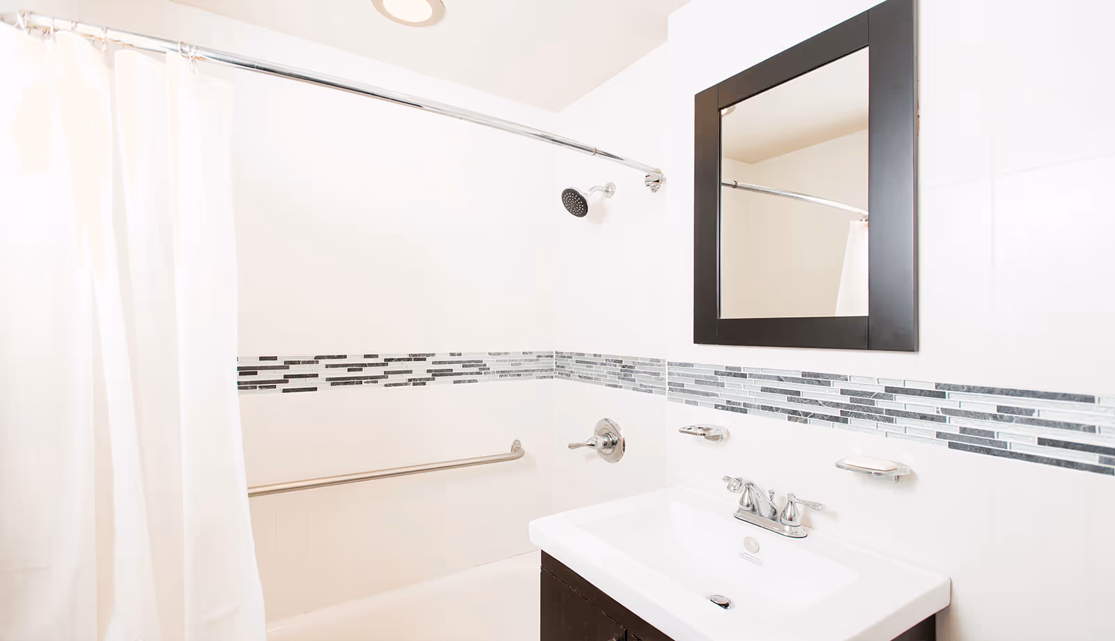 A clean, modern bathroom featuring a white bathtub with a curved shower curtain rod and white shower curtain. The wall has a decorative horizontal strip of black, gray, and white tiles. There is a silver showerhead and a grab bar on the wall. A white sink with a silver faucet is below a rectangular mirror with a dark frame.