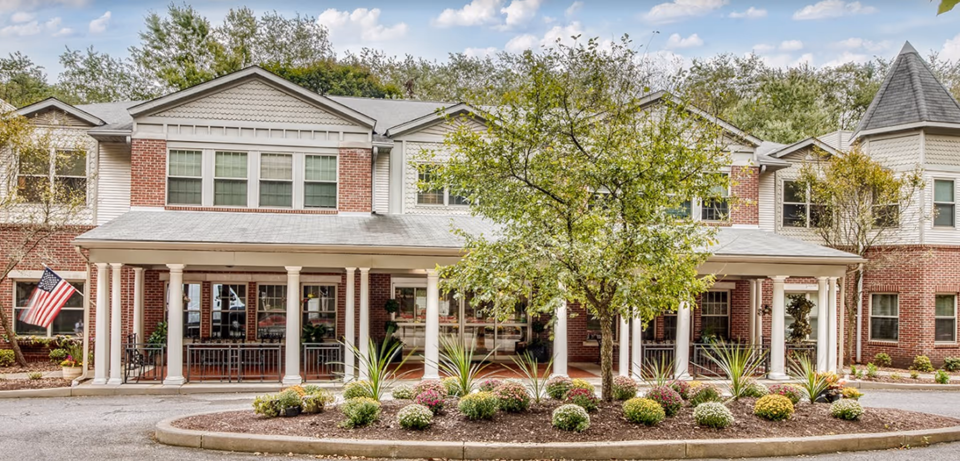 Front exterior view of a two-story senior living facility with a covered porch supported by white columns. The building has a combination of red brick and light-colored siding, multiple windows, and a small landscaped area with bushes and a tree in front. An American flag is displayed on the left side of the porch.