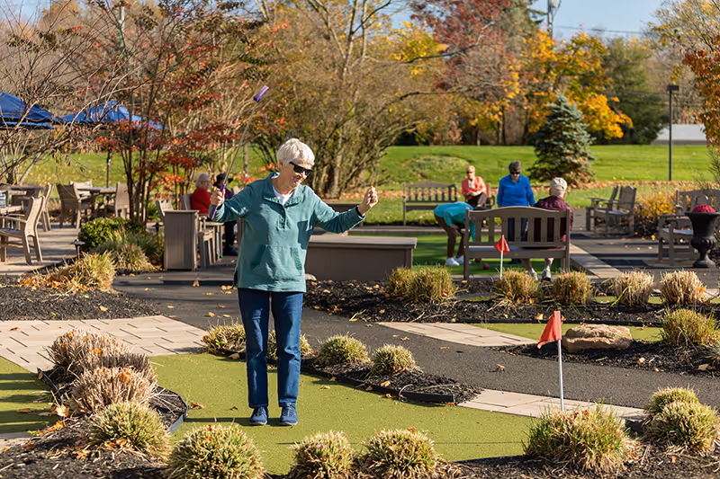 An elderly woman playing miniature golf outdoors on a sunny day, surrounded by bushes and trees with autumn foliage. Several other elderly people are sitting on benches and socializing in the background.