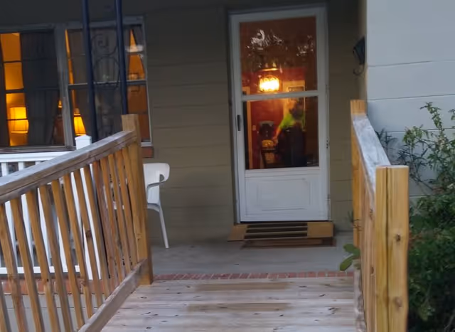 Exterior porch with wooden railing leading to a white door and a chair on the deck.