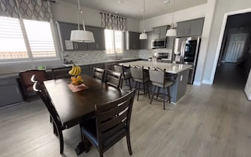 Open modern kitchen and dining area with a dark wood table, island with barstools, gray cabinets, and pendant lights.
