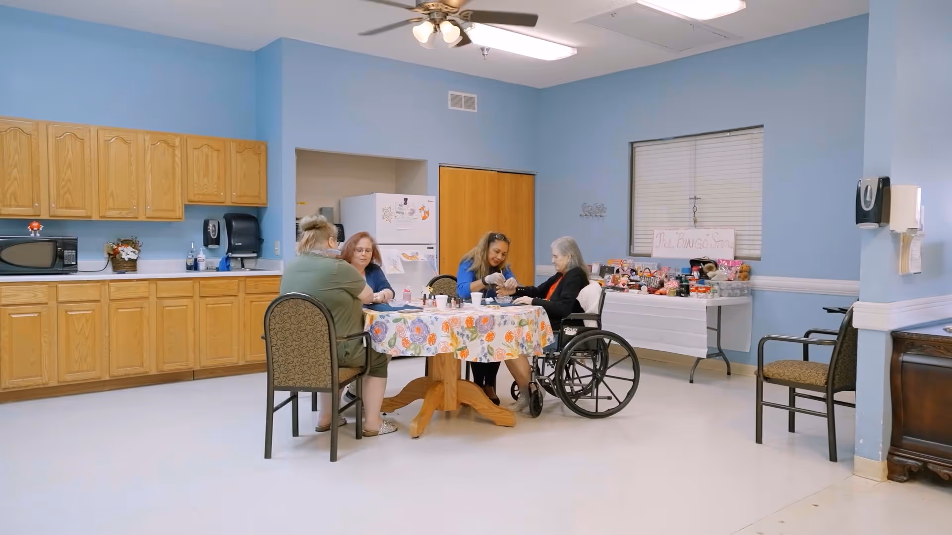 Four women seated around a floral-covered table in a light-blue communal room with kitchen cabinets and a snack table, one woman in a wheelchair.