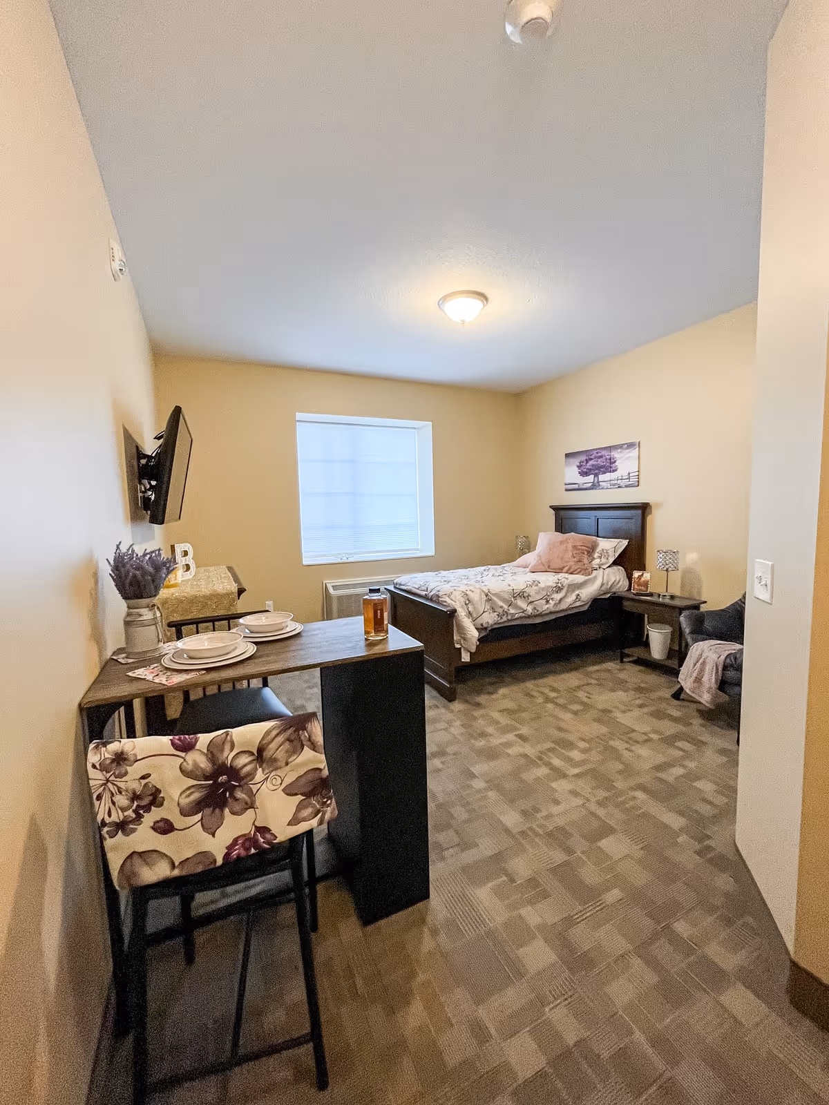 A small, cozy bedroom with a single bed against the far wall, decorated with a floral comforter and a pink pillow. There is a small table with two chairs near the entrance, set with plates and bowls. A flat-screen TV is mounted on the left wall. The room has beige walls, a window with a closed blind, and carpeted flooring. A purple-themed picture hangs above the bed, and a small armchair with a pink throw is placed near the bed.