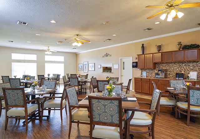 A bright dining room with multiple wooden tables and chairs arranged neatly. Each table has a small floral centerpiece and place settings. The room features large windows allowing natural light, ceiling fans with lights, wooden flooring, and a counter area with cabinets and a coffee machine.