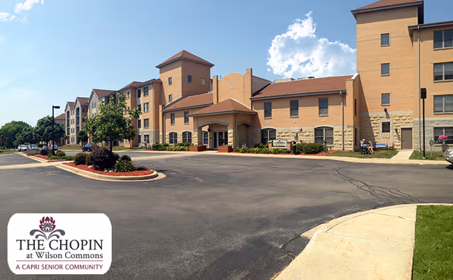 Exterior view of a multi-story senior living facility building with beige and light brown walls, a driveway, landscaped greenery, and a clear blue sky.