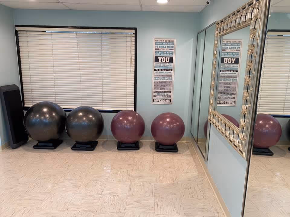 A small exercise room with four large exercise balls placed on black stands against a light blue wall. There is a window with closed blinds above the balls, a motivational poster on the wall, and a large decorative mirror reflecting part of the room.