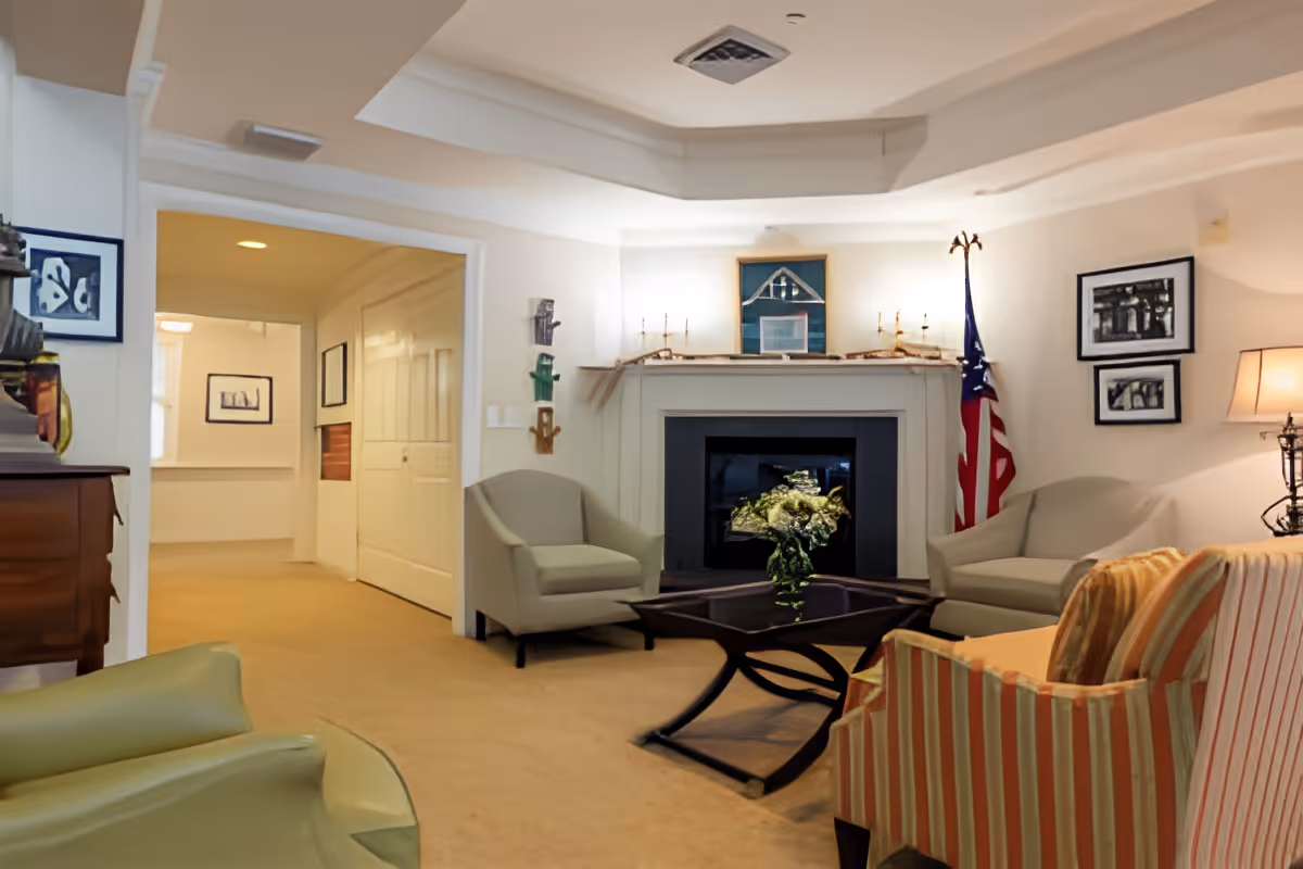 A cozy living room with beige carpet, a fireplace with decorative items on the mantel, an American flag in the corner, and several armchairs arranged around a dark wooden coffee table with a flower arrangement. The room has soft lighting and framed pictures on the walls.