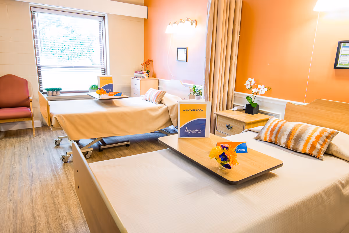 A bright and clean room with two hospital-style beds, each with a wooden headboard and a beige bedspread. Both beds have overbed tables with flowers and welcome books on them. The room has an orange accent wall, a window with blinds, a wooden nightstand with a white orchid, and a pink upholstered chair near the window.