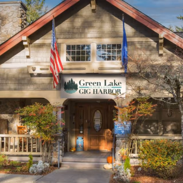 Front entrance of Green Lake Senior Living Gig Harbor with a sign above wooden double doors, flanked by flags and landscaping.