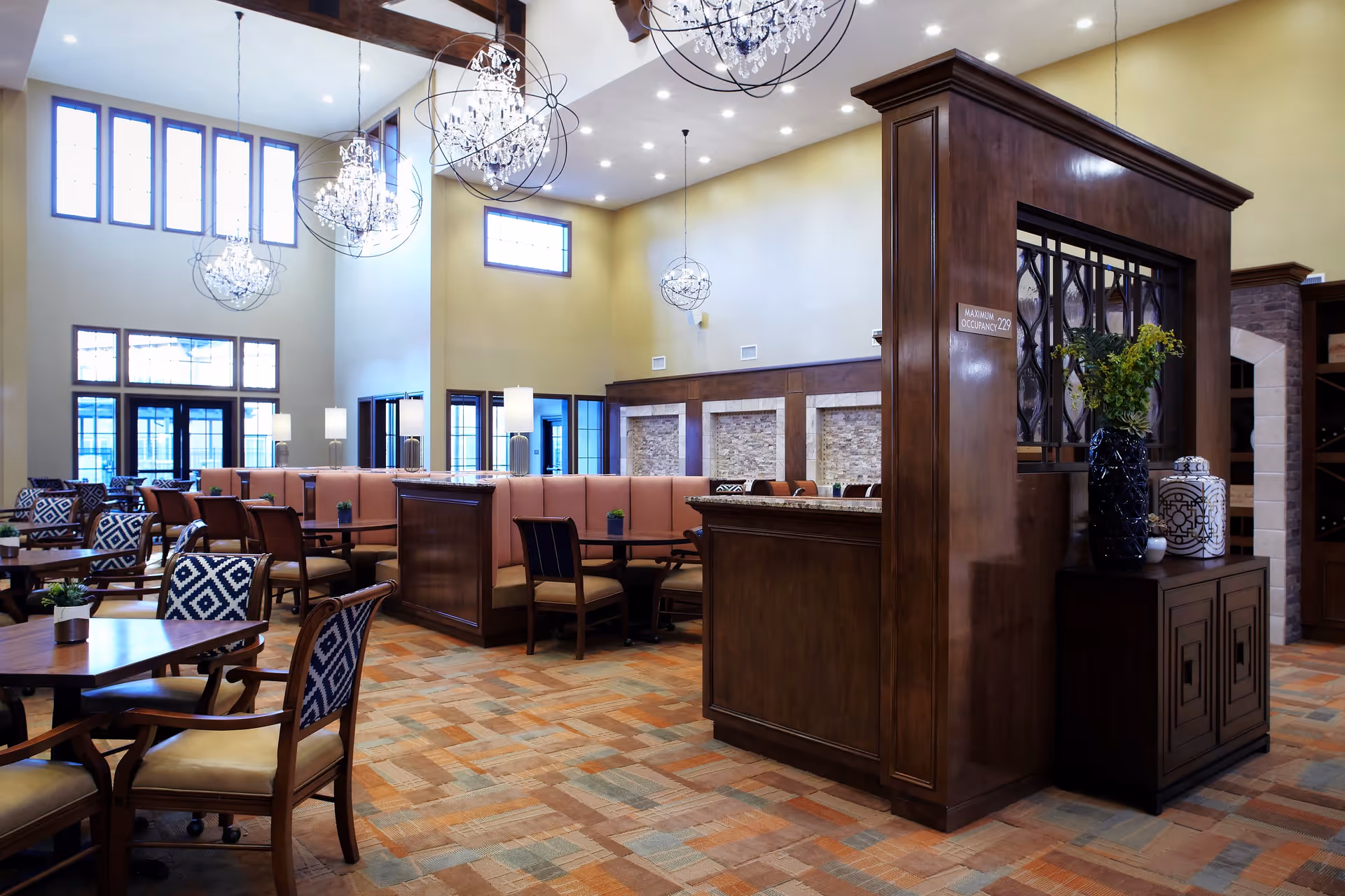 A spacious and well-lit dining area with multiple tables and chairs, featuring patterned upholstery on some chairs. The room has high ceilings with large windows, elegant chandeliers, and wooden beams. There is a wooden partition with decorative elements and a vase with flowers, and the floor is covered with a patterned carpet.
