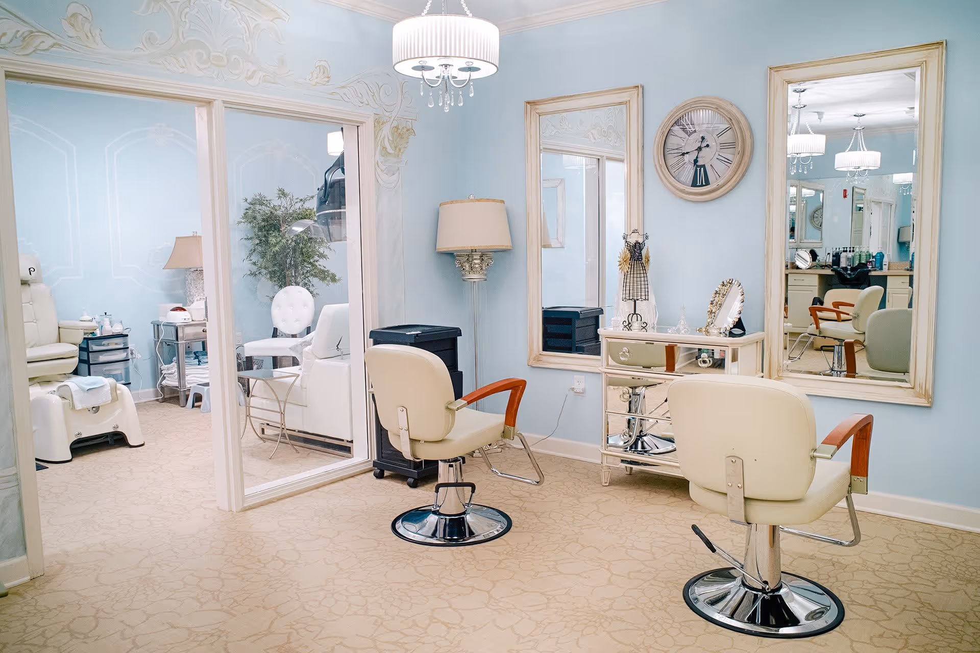 A bright and elegant salon area with two cream-colored styling chairs facing large mirrors mounted on a light blue wall. The room features a chandelier, a floor lamp, a mirrored dresser with decorative items, and a separate adjacent room with a pedicure chair and other salon equipment.