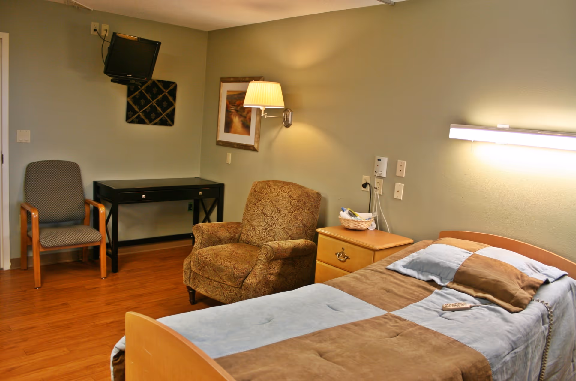 A bedroom in a senior living facility with a single bed covered in a blue and brown blanket, a matching pillow, and a remote control on the bed. Next to the bed is a wooden nightstand with a basket containing toiletries. There is a patterned armchair and a wooden chair with armrests near a black desk. A wall-mounted TV is positioned in the corner above the desk. The walls are painted light green, and there is a framed picture and a wall lamp above the armchair. The floor is wooden.
