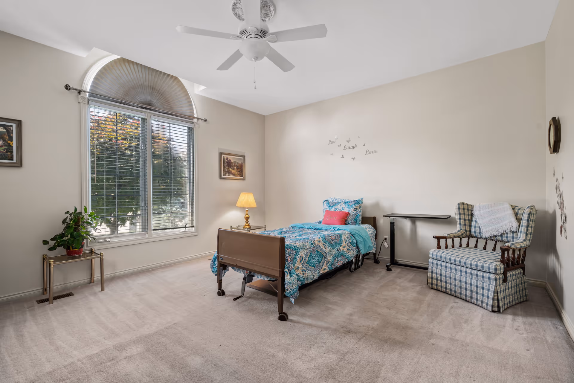 A bright bedroom with a single bed covered in a blue patterned comforter and a pink pillow. There is a small side table with a lamp next to the bed, a plaid upholstered armchair with a white throw draped over it, and a small overbed table. The room has beige walls, a large window with blinds and a decorative fan-shaped curtain, a ceiling fan, and a potted plant on a small stand near the window. Wall decor includes framed pictures and a 'Live Laugh Love' decal with butterfly accents.