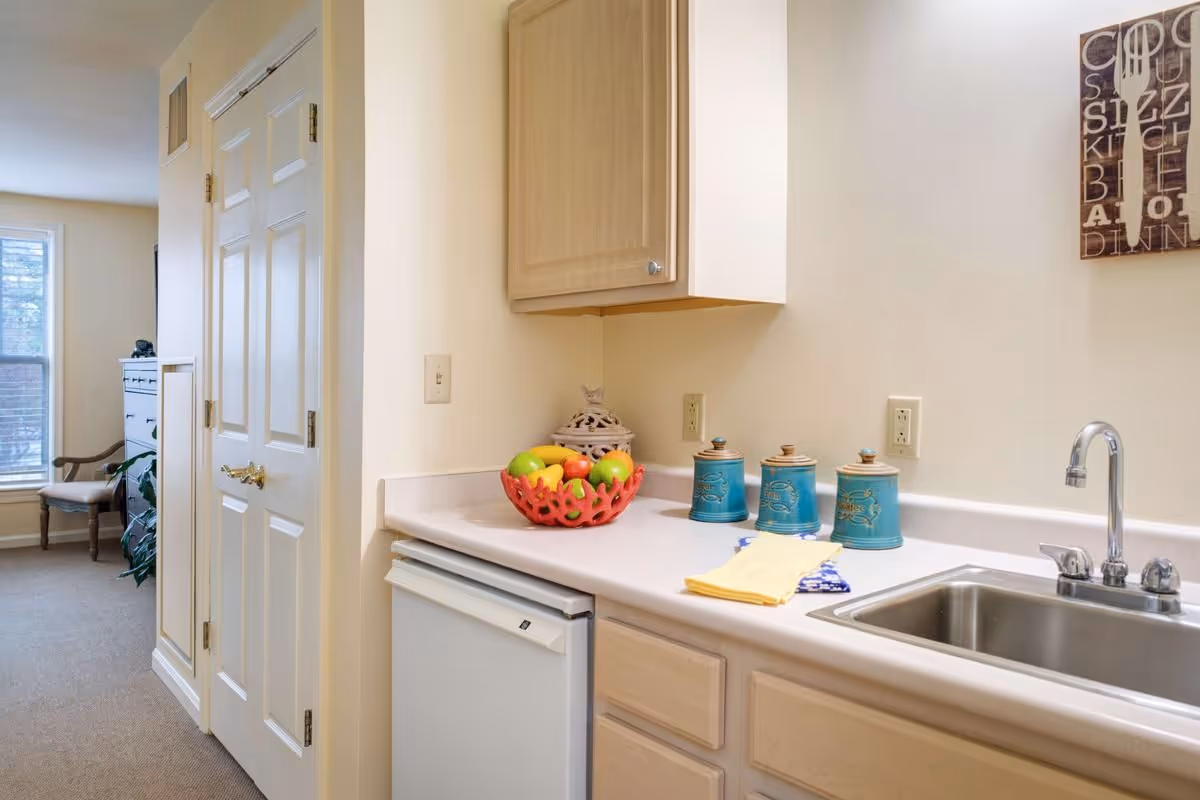 Compact kitchenette featuring a sink, countertop with a fruit bowl and teal canisters, light wood cabinets, and a small refrigerator.