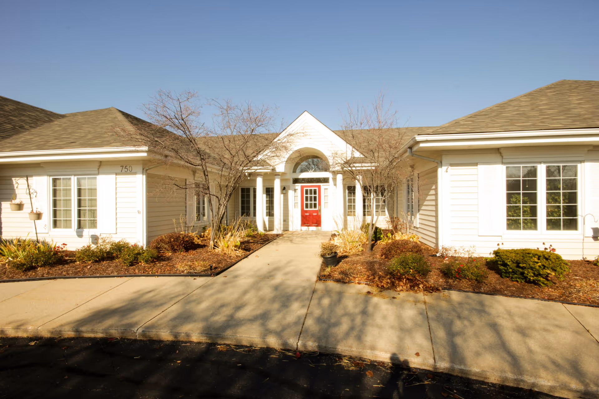 Exterior front view of a single-story building with a red door entrance, white siding, multiple windows, and a small garden with shrubs and leafless trees along the walkway.