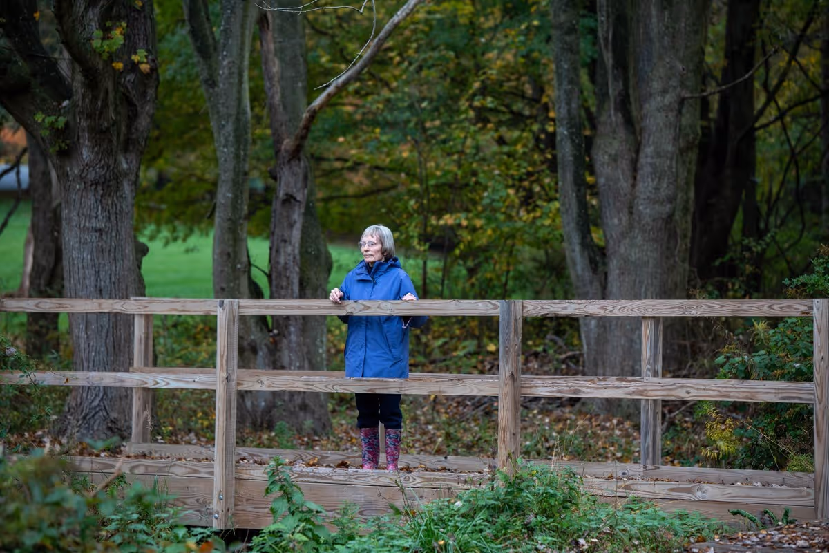 An elderly woman wearing a blue coat and colorful boots stands on a wooden bridge surrounded by trees and greenery in an outdoor setting.