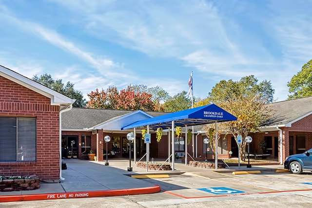 Exterior view of a single-story brick building with a blue canopy entrance labeled 'Brookdale Champion Oaks'. There are handicap parking spaces and a red curb marked 'Fire Lane No Parking'. Trees and a clear sky are visible in the background.