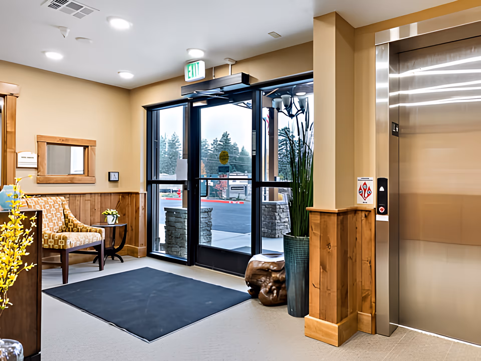 Bright entrance lobby with a seating area, glass double doors, potted plants, and an elevator.
