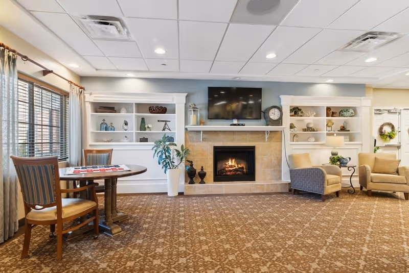 A cozy living room area with a lit fireplace in the center, a flat-screen TV mounted above it, and built-in white shelves on either side displaying decorative items. There are two beige armchairs with a small side table between them on the right, and a round wooden table with two chairs on the left near a window with curtains. The room has a patterned carpet and a ceiling with recessed lighting.