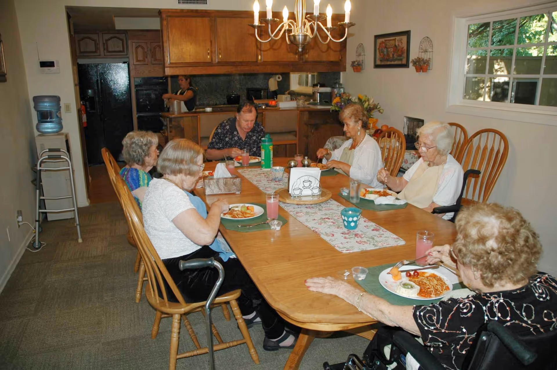 Several older adults sit around a long dining table eating a meal in a dining room with the kitchen visible in the background.