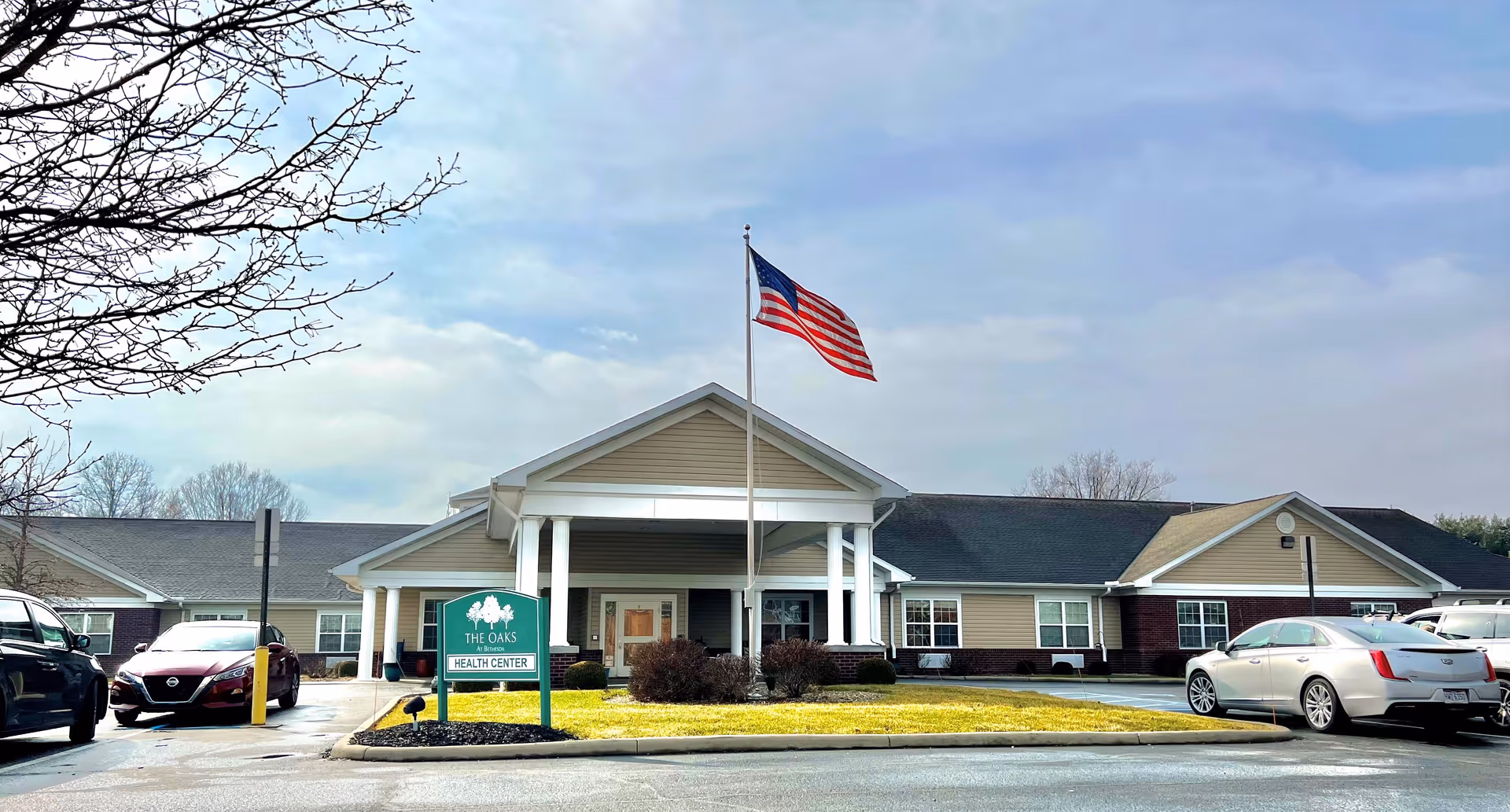 Front exterior of The Oaks at Bethesda health center with an American flag on a pole and cars parked in the driveway.