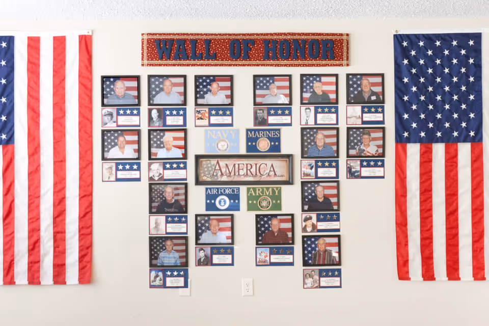A wall display titled 'Wall of Honor' featuring framed photographs of veterans in front of American flags, along with plaques representing different military branches including Navy, Marines, Air Force, and Army. Two large American flags hang on either side of the display.