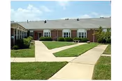 Courtyard with intersecting concrete walkways and grass in front of a single-story brick senior living building under a blue sky.
