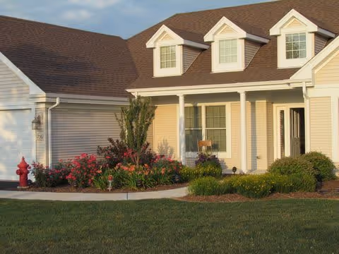 Exterior view of a single-story residential building with beige siding, a brown shingled roof, three dormer windows, a white front porch with columns, and a landscaped garden with flowers and shrubs in front. A red fire hydrant is visible on the left side near the garden.