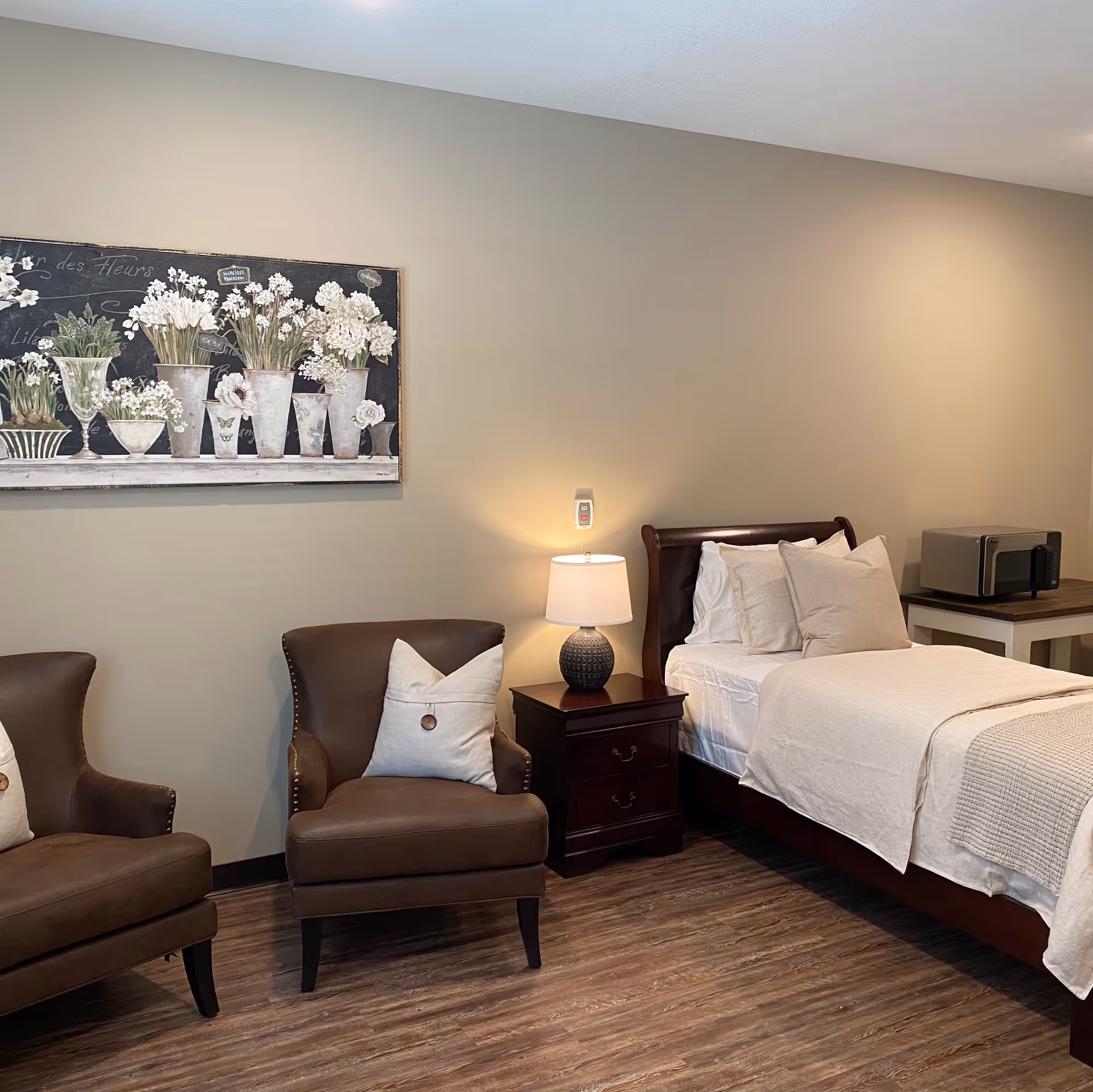 A cozy bedroom in an assisted living facility featuring a single bed with white bedding and multiple pillows, a wooden nightstand with a lamp, two brown leather armchairs with white pillows, a microwave on a small table, and a floral painting on the beige wall.