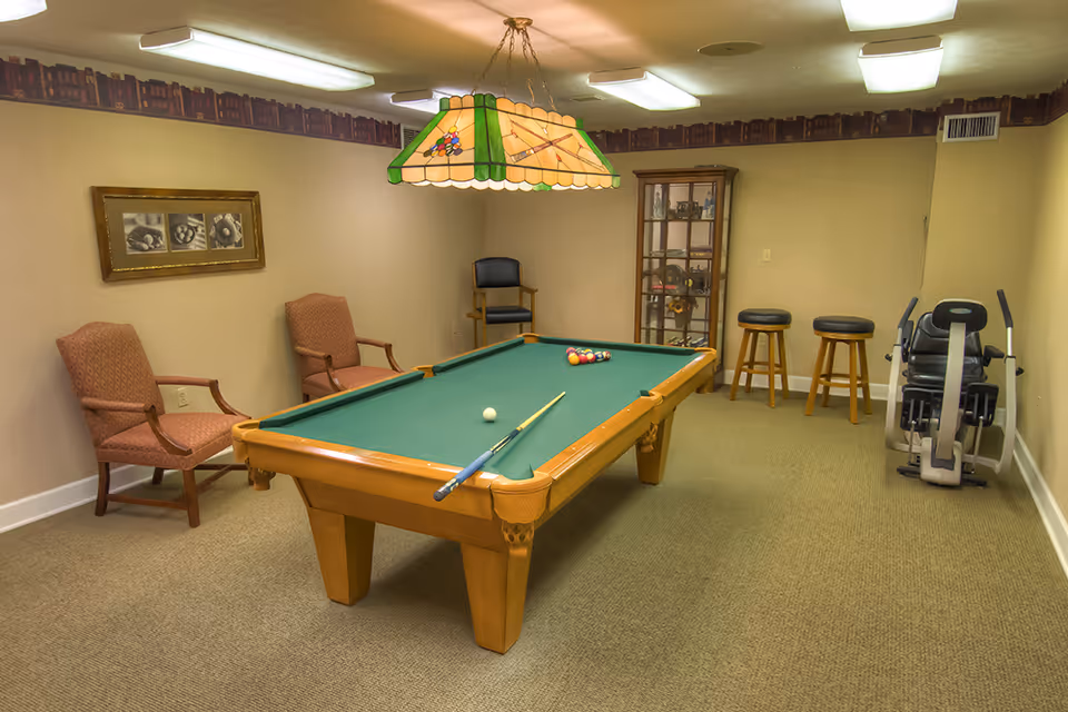 A recreational room with a pool table in the center, two pool cues resting on the table, and a rack of billiard balls. The room has beige walls and carpet, two upholstered armchairs, a wooden display cabinet, two bar stools, and an exercise machine. A stained glass billiard light fixture hangs above the pool table.