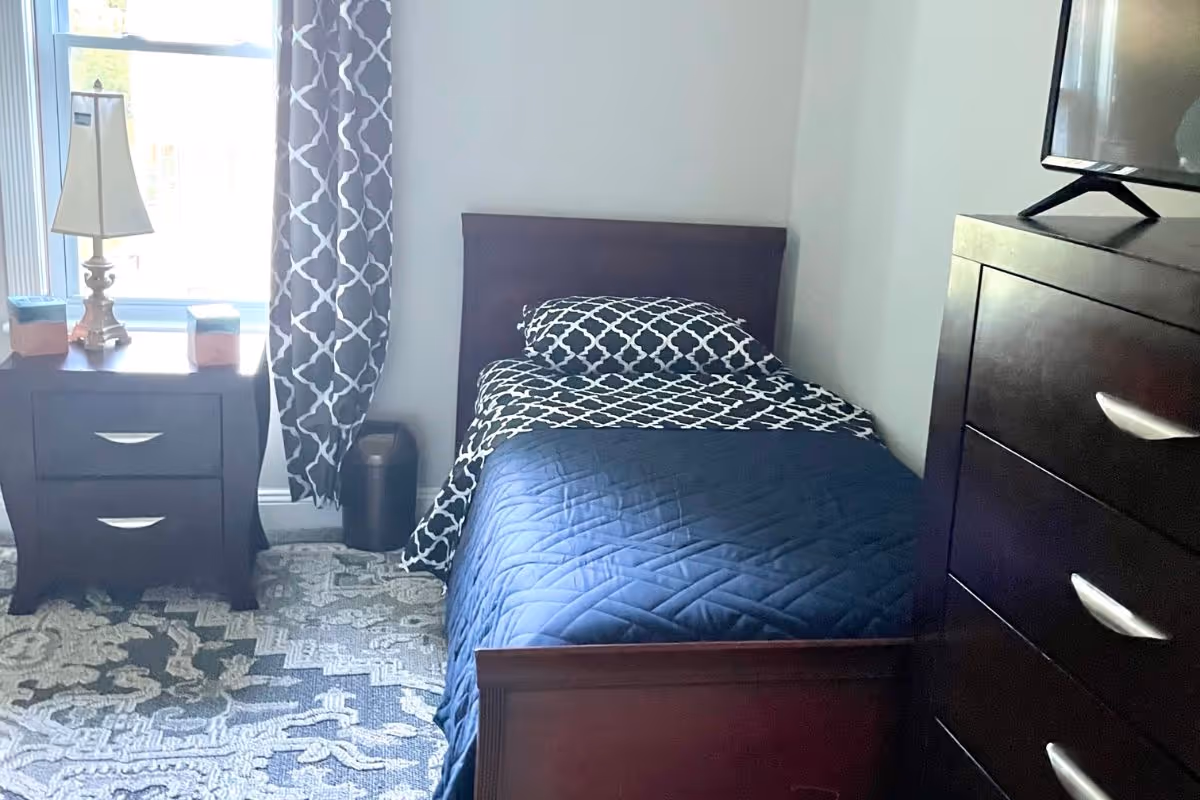 A small bedroom with a single bed covered with a dark blue quilt and a black and white patterned pillow and blanket. Next to the bed is a wooden nightstand with two drawers, a lamp, and two small decorative boxes. A window with patterned curtains is behind the nightstand. On the right side of the image, there is a dark wooden dresser with silver handles and a TV on top.