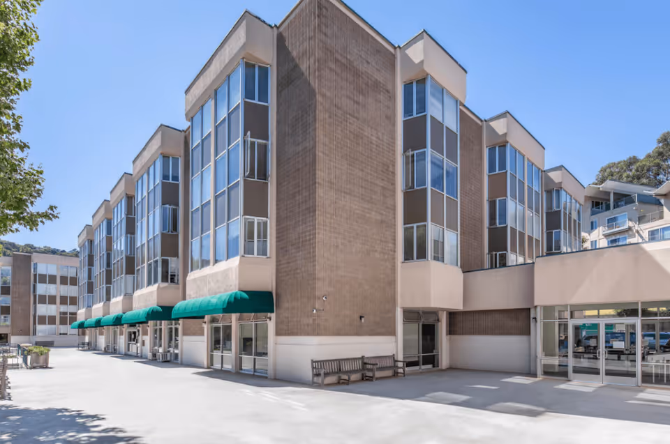 Exterior front view of a multi-story senior living facility with large windows, green awnings, and a paved courtyard.