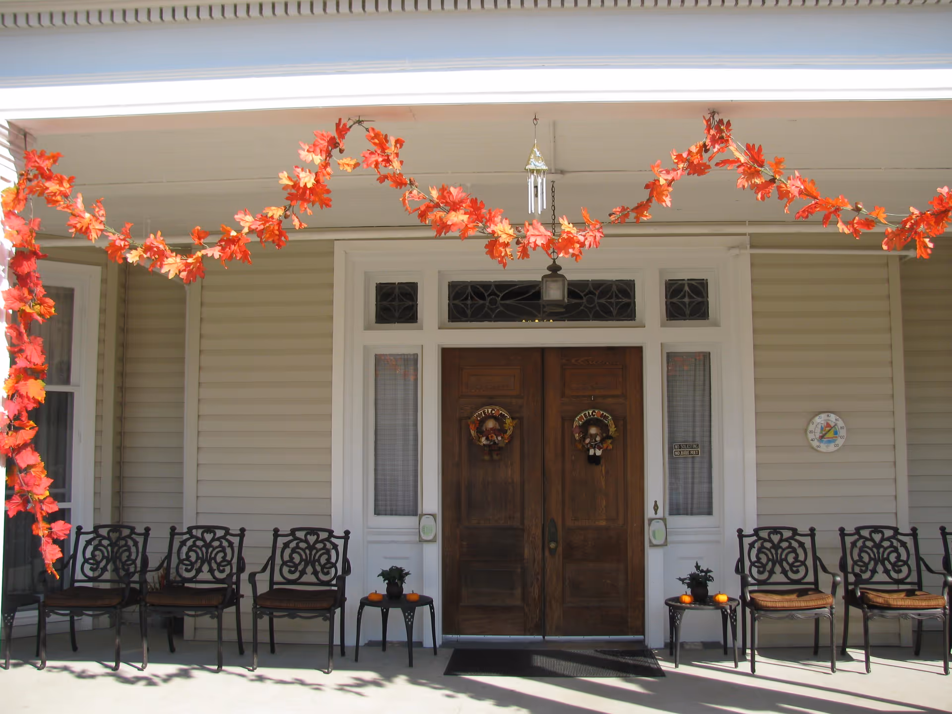 Front porch of a building with double wooden doors decorated with autumn wreaths. There are six ornate metal chairs with cushions arranged on either side of the doors, and two small tables with pumpkins and potted plants. Orange and red autumn leaf garlands hang across the porch ceiling. A hanging lantern and a wind chime are above the doors, and a round thermometer is mounted on the wall to the right.