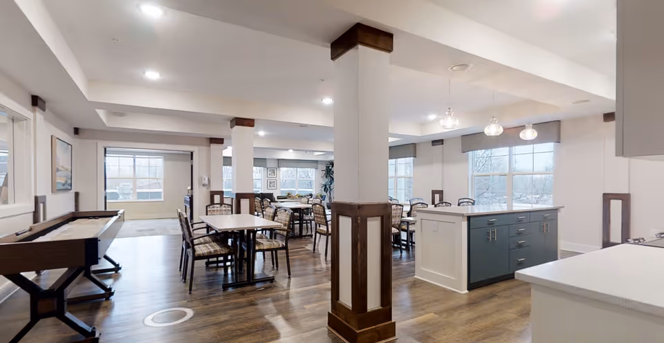 Open communal dining and activity room with tables and chairs, a kitchen island, and a shuffleboard table.
