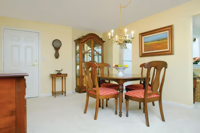 A dining area in a senior living facility with a wooden dining table and four chairs with red cushioned seats. A white fruit bowl with assorted fruit is on the table. Behind the table is a wooden china cabinet with glass doors. A wall clock and a small side table are near a white door. A framed landscape painting hangs on the wall, and a chandelier with lit bulbs hangs above the dining table. The walls are painted light yellow and the floor is carpeted in white.