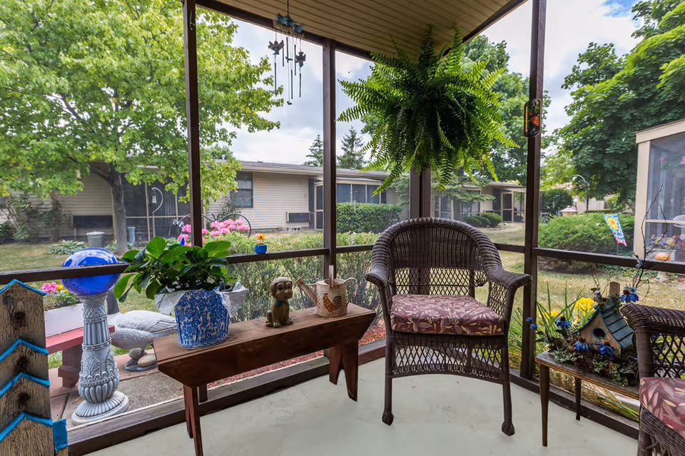 A screened-in porch area with wicker chairs featuring floral cushions, a wooden bench with potted plants and decorative items, and a hanging fern plant. Outside the porch, there is a garden with bushes, trees, and a building in the background.